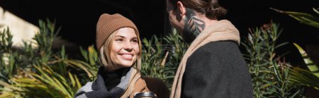 Foto de Mujer rubia feliz en sombrero beige mirando novio tatuado en abrigo mientras sostiene el café para llevar, pancarta - Imagen libre de derechos