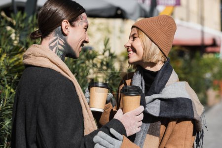 Photo for Side view of happy blonde woman in beige hat looking at tattooed boyfriend in coat while holding coffee to go - Royalty Free Image