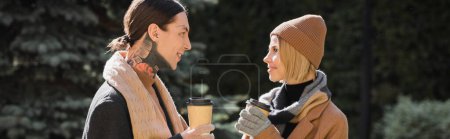 Téléchargez les photos : Vue latérale de femme gaie en chapeau beige regardant son petit ami tatoué en manteau tout en tenant du café à emporter, bannière - en image libre de droit