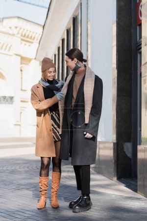 full length of happy woman in hat adjusting scarf of man in coat 