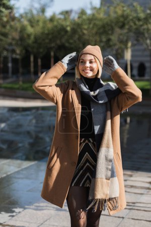 mujer rubia feliz en abrigo y guantes ajustando sombrero de pie sonriendo fuera 