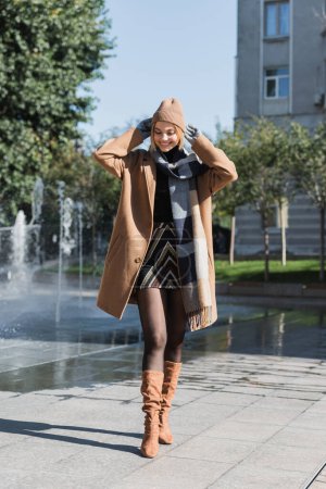 full length of happy blonde woman in coat and gloves adjusting hat standing smiling outside 