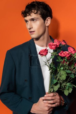 Stylish brunette man in blazer holding roses on orange background 