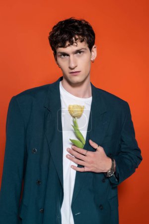 Trendy brunette man holding tulip behind jacket on red background 