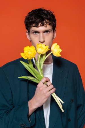 Trendy brunette man in blue blazer holding yellow tulips isolated on red 
