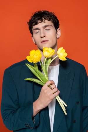 Brunette man in jacket closing eyes and holding yellow tulips isolated on red 