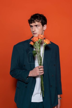 Téléchargez les photos : Trendy young man in blazer holding orange roses for 8 march on red background - en image libre de droit