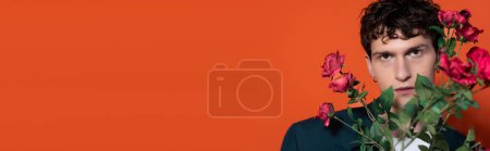 Photo for Curly brunette man looking at camera near roses on red background, banner - Royalty Free Image
