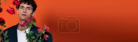 Stylish curly model looking at camera near roses on red background, banner 