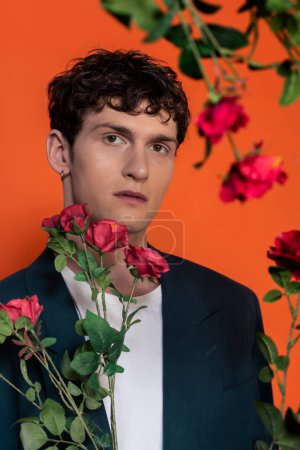 Hombre rizado en camiseta y chaqueta mirando a la cámara cerca de rosas aisladas en rojo 