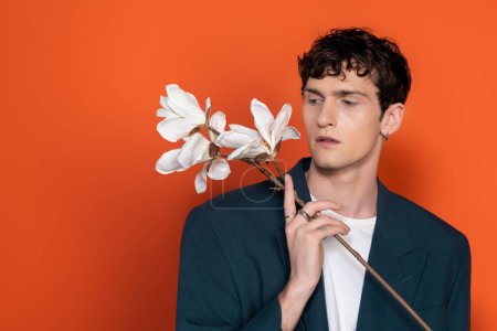 Stylish brunette man looking at magnolia flowers on orange background