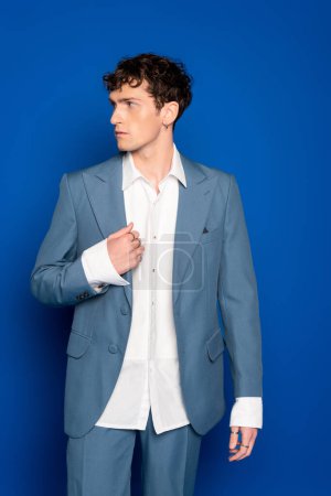 Joven hombre de moda en traje y camisa mirando hacia otro lado sobre fondo azul 