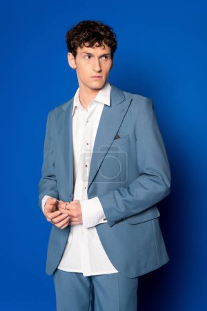Hombre morena de moda en traje y camisa blanca mirando hacia otro lado sobre fondo azul 