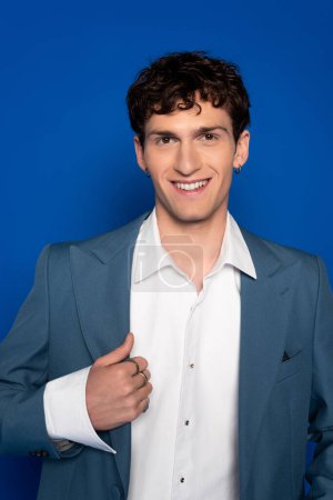 Portrait of smiling and curly model in blazer looking at camera on blue background 