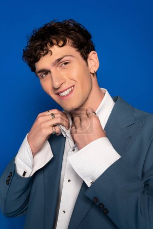 Photo for Portrait of cheerful curly man in jacket adjusting shirt isolated on blue - Royalty Free Image