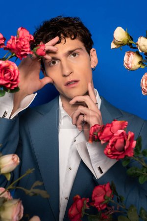 Portrait of stylish curly model in shirt and jacket touching face near roses on blue background 