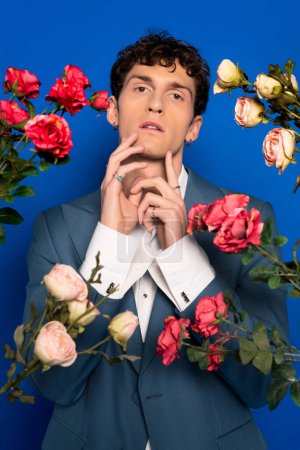 Curly man in stylish outfit touching chin near roses on blue background 
