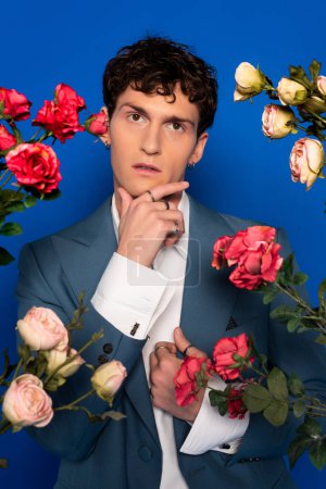 Lockiger Mann in stylischem Outfit, der das Kinn in der Nähe blühender Blumen auf blauem Hintergrund berührt 