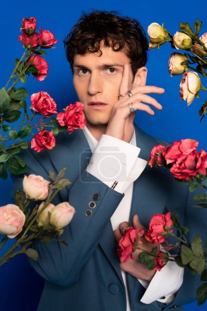 Stilvoller Mann in Hemd und Jacke posiert in der Nähe von Rosen auf blauem Hintergrund 