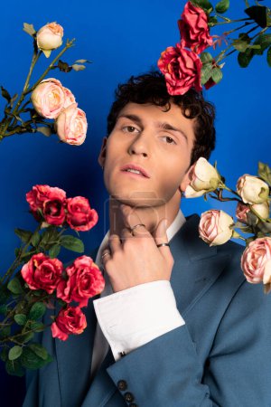 Trendiger junger Mann in Jacke posiert in der Nähe von Rosen auf blauem Hintergrund 