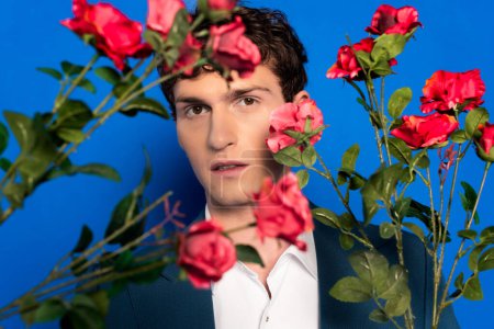Jeune homme en veste regardant la caméra près de fleurs roses floues sur fond bleu 