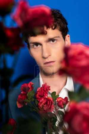 Trendy young man looking at camera near flowers on blue background 