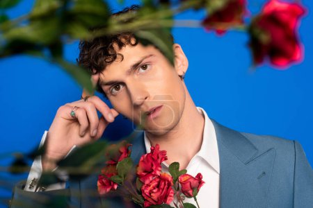 Lockiger und stilvoller junger Mann blickt in die Kamera in der Nähe von Rosen auf blauem Hintergrund 