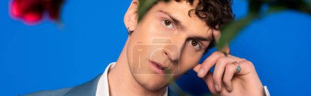 Trendy and curly man looking at camera near blurred flower on blue background, banner 
