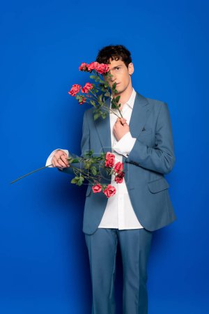 Trendy model in suit holding flowers near face on blue background 