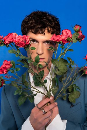 Curly model in jacket holding roses near face on blue background 