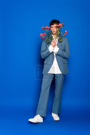 Téléchargez les photos : Full length of stylish man in suit holding flowers on blue background - en image libre de droit