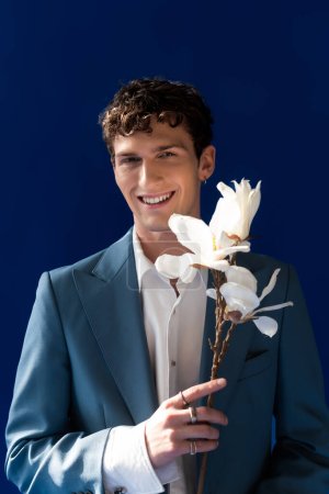 Hombre positivo en traje elegante sosteniendo rama magnolia aislado en azul marino 