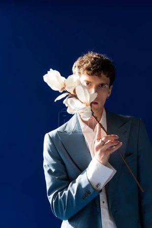Stilvoller Mann im Blazer mit Magnolienblüten in Gesichtsnähe isoliert auf marineblau 