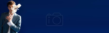 Foto de Curly man in stylish outfit holding magnolia flower isolated on navy blue, banner - Imagen libre de derechos