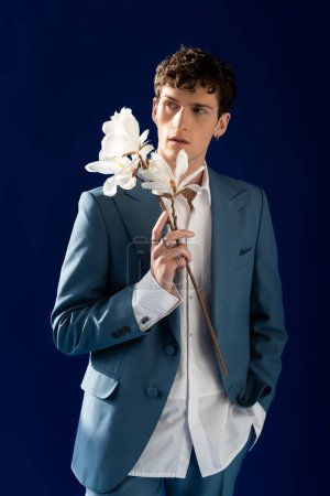 Téléchargez les photos : Fashionable model in suit holding magnolia flowers isolated on navy blue with sunlight - en image libre de droit