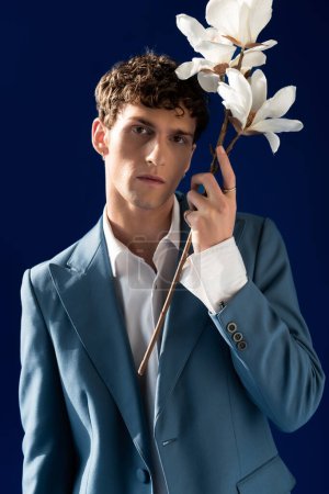 Retrato de un joven elegante con chaqueta que sostiene flores de magnolia aisladas en azul marino 