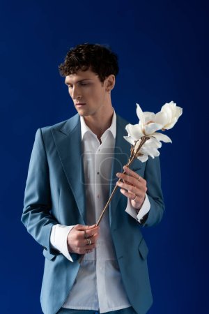 Hombre morena en chaqueta elegante y camisa que sostiene magnolia rama aislado en azul marino 
