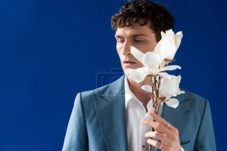Portrait of stylish curly man holding magnolia branch isolated on blue 