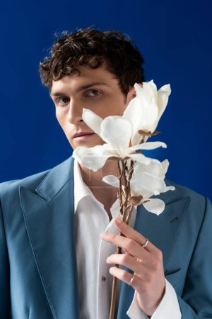 Trendy young man in jacket and shirt holding magnolia flowers isolated on blue 