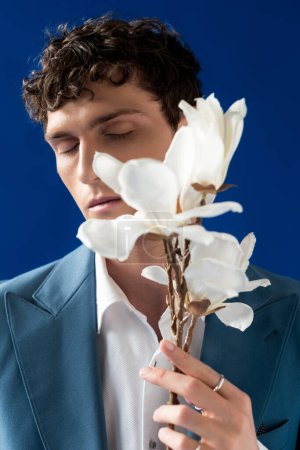 Photo for Portrait of curly young man in jacket holding blurred magnolia branch isolated on blue - Royalty Free Image