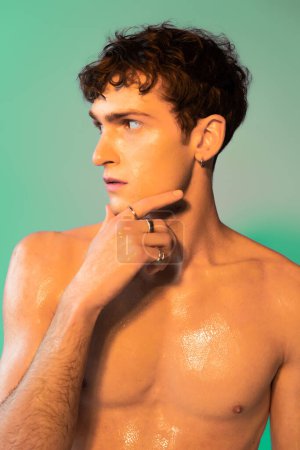 Shirtless model with oil on body touching chin on green background 