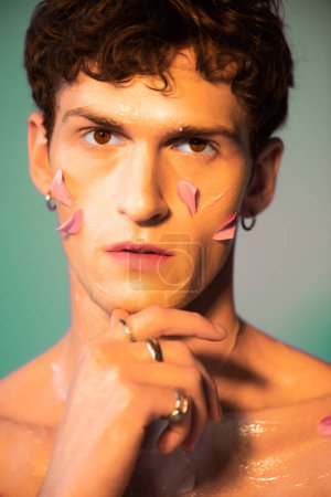 Shirtless brunette model with petals on face looking at camera on colorful background 