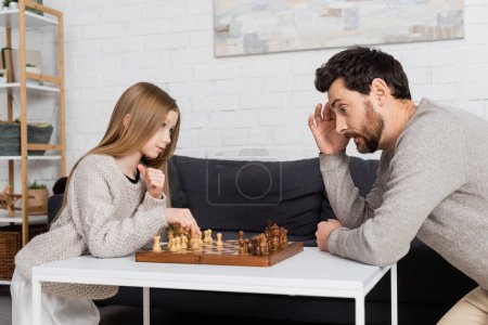 thoughtful man touching head while playing chess with preteen daughter in living room
