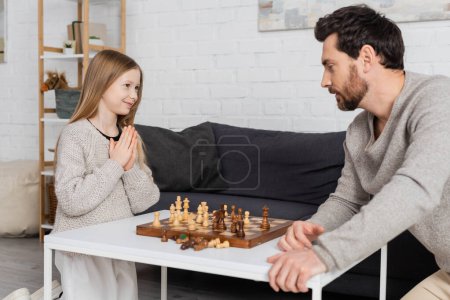 smiling girl showing please gesture while playing chess with father at home