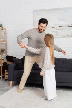full length of joyful bearded man teaching daughter to dance near sofa in modern living room