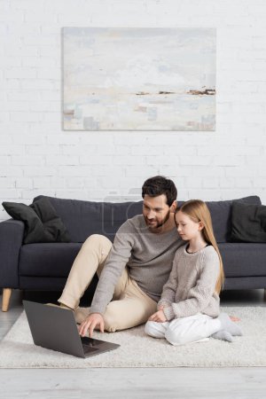 Téléchargez les photos : Preteen girl sitting near father using laptop on floor carpet in living room - en image libre de droit