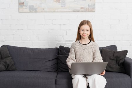 Photo for Pleased girl sitting on couch with laptop and smiling at camera - Royalty Free Image