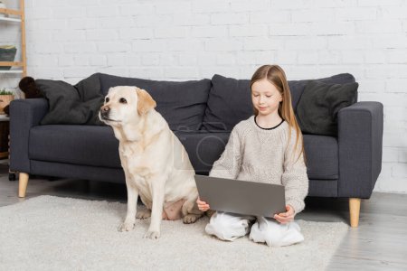 positive preteen girl sitting on floor carpet near labrador dog and looking at laptop