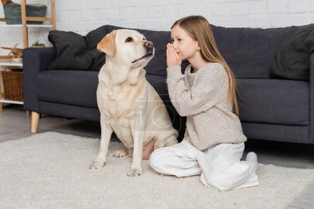 preteen girl sitting on floor near couch and telling secret to labrador dog