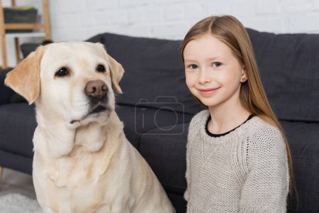 Téléchargez les photos : Joyeuse fille souriant à la caméra près du chien labrador dans le salon - en image libre de droit